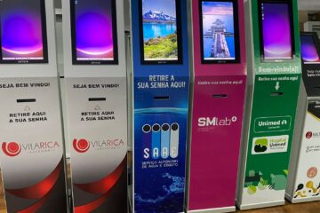 Colorful self-service kiosk row with screens and Portuguese prompts for passwords and welcome messages (Seja bem vindo!, Retire aqui a sua senha) in a bank/clinic setting.
